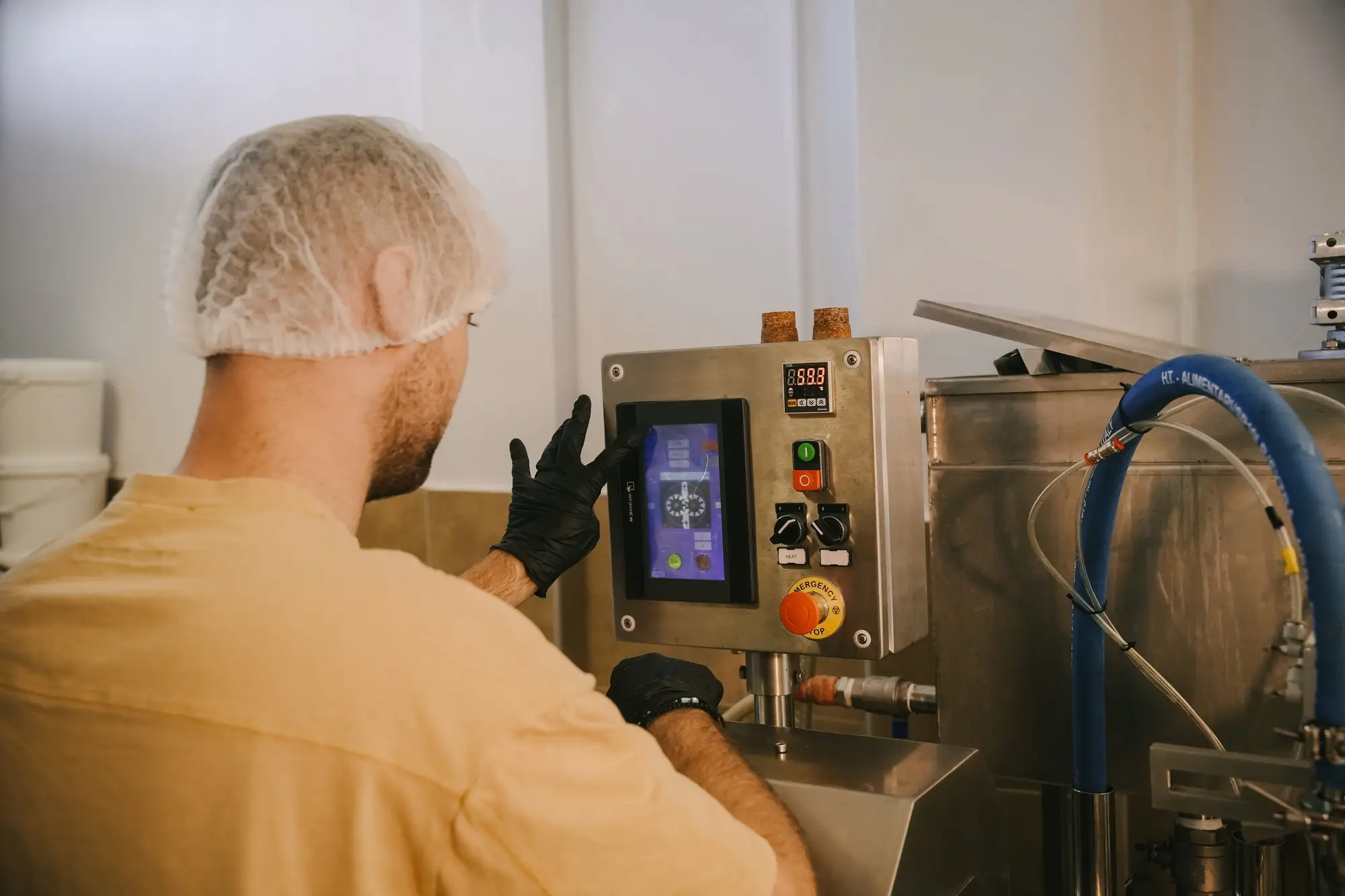 Man wearing a hairnet and black gloves operating a touchscreen control panel on industrial machinery.