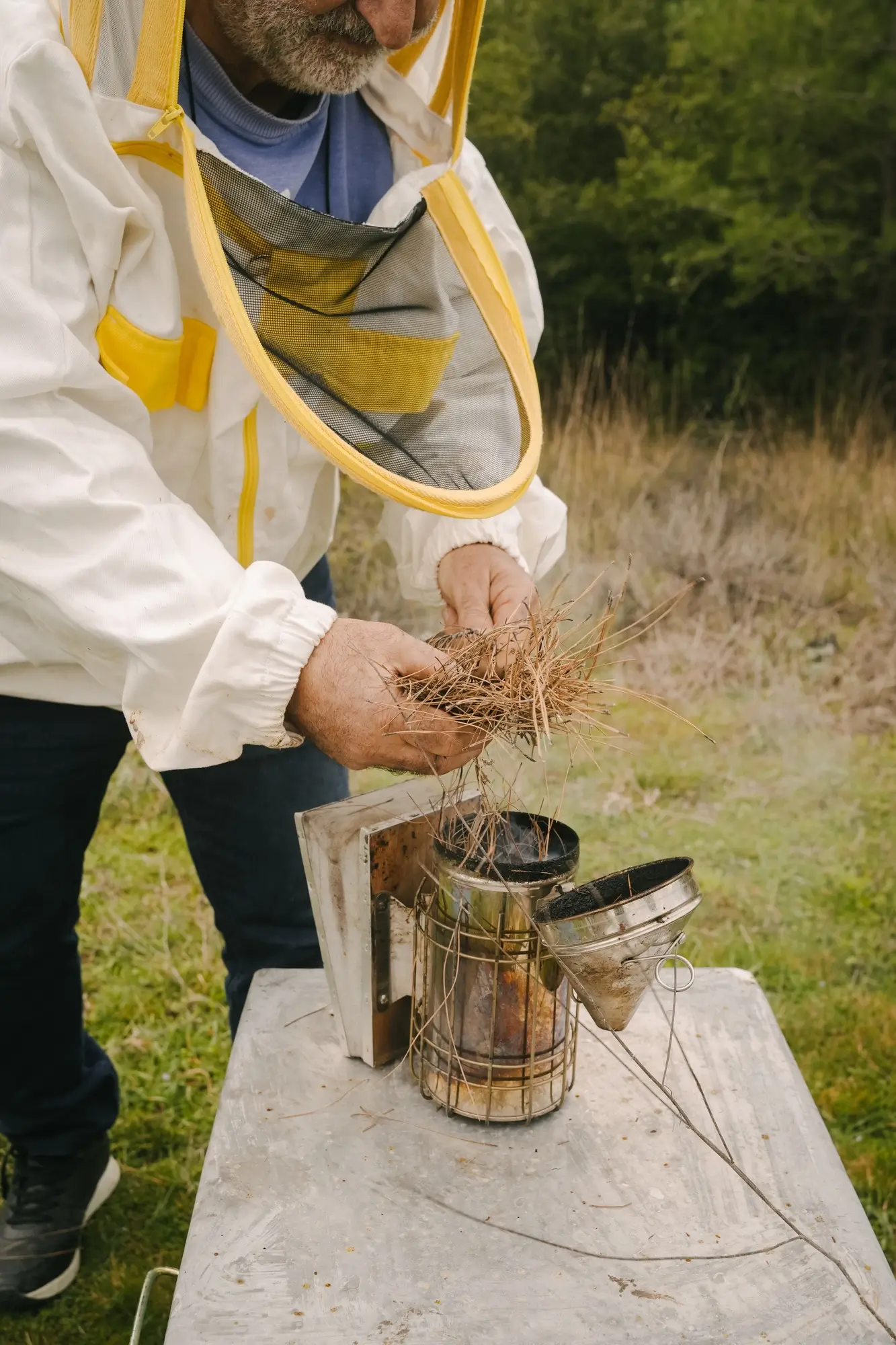 Beekeeper in protective suit preparing a smoker with dry grass outdoors.