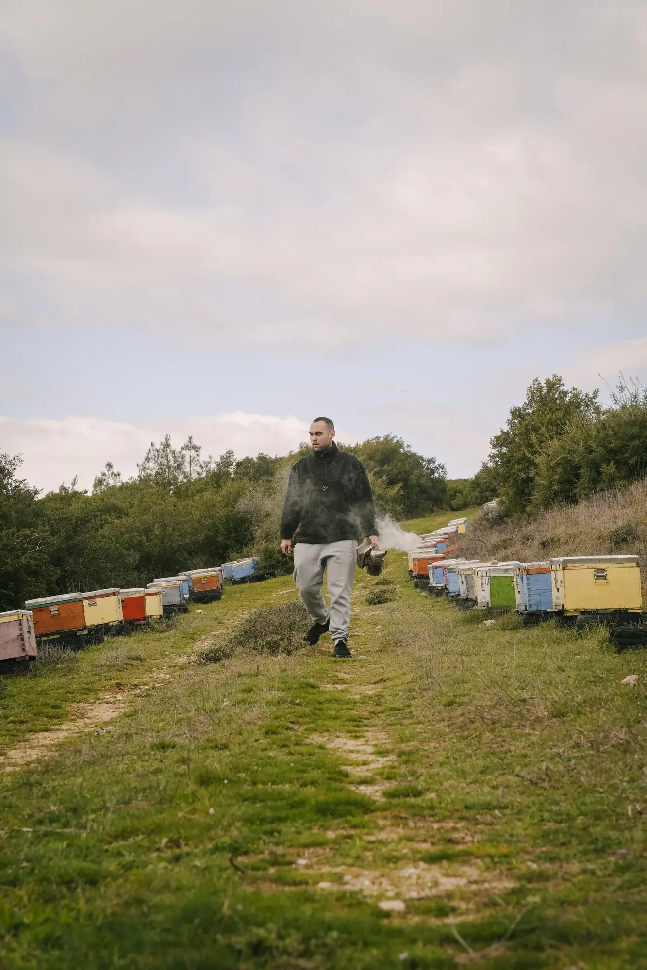 Man walking on a grassy path between colorful beehives holding a smoking bee smoker.