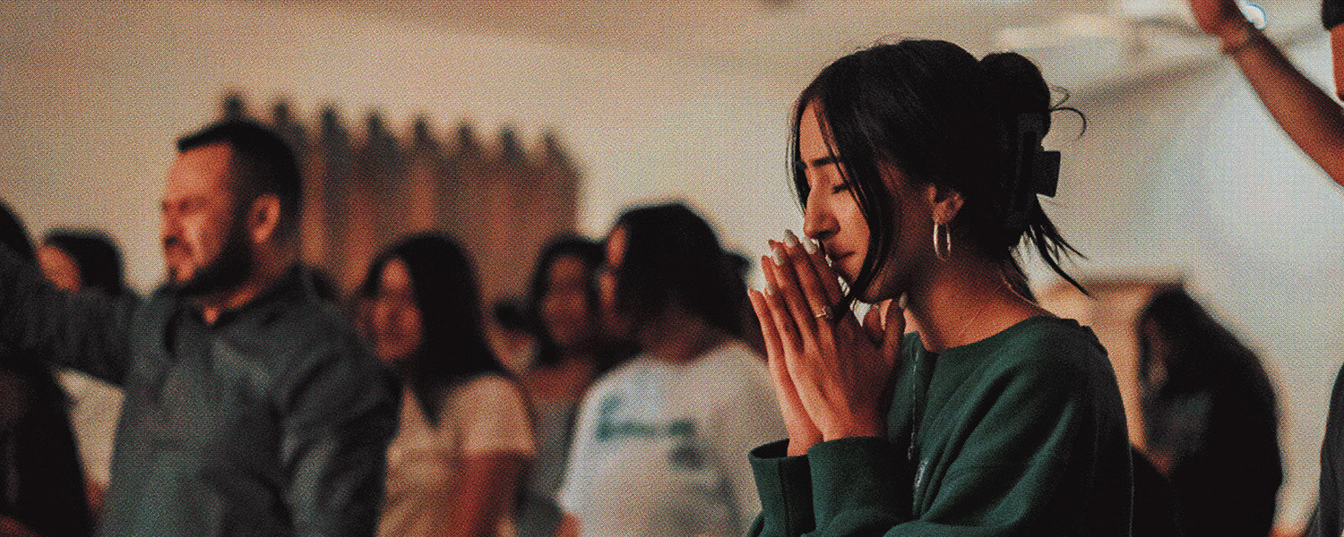 Woman with eyes closed praying with hands together among a group of people in a softly lit room.