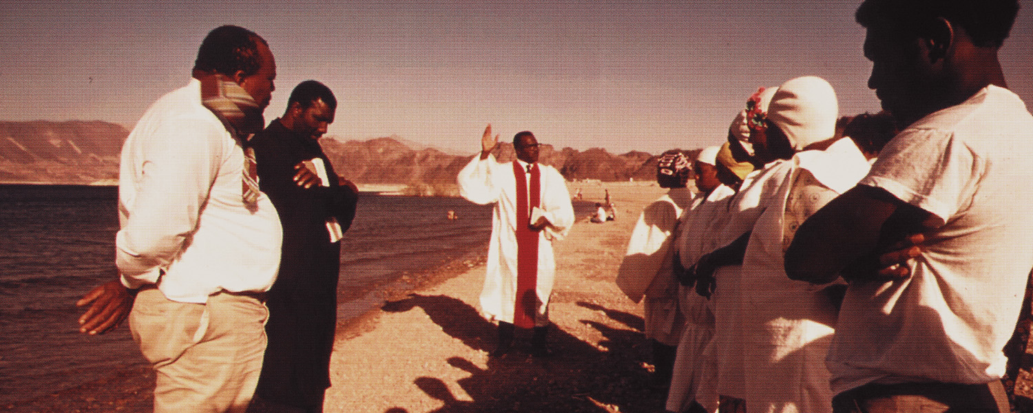 A group of people standing by a lake; one man in white robes is speaking or praying to the group.