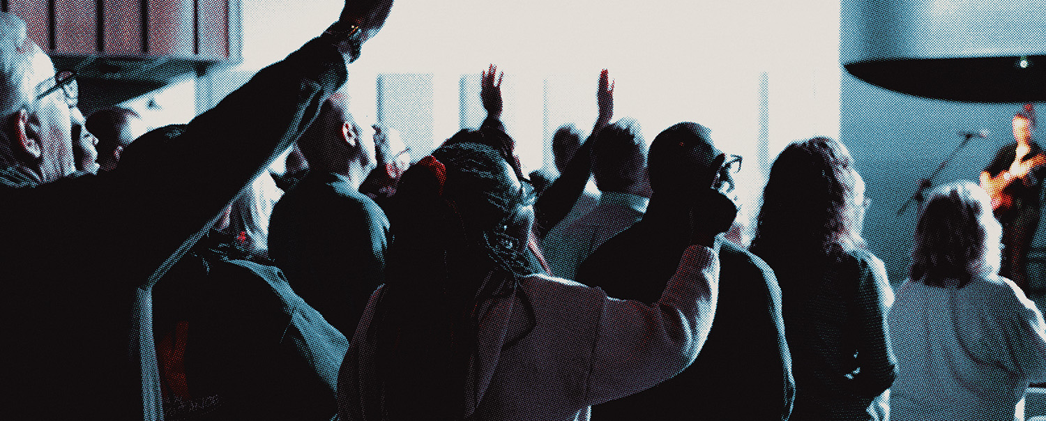 Group of people standing indoors with hands raised, facing a musician playing guitar on stage.