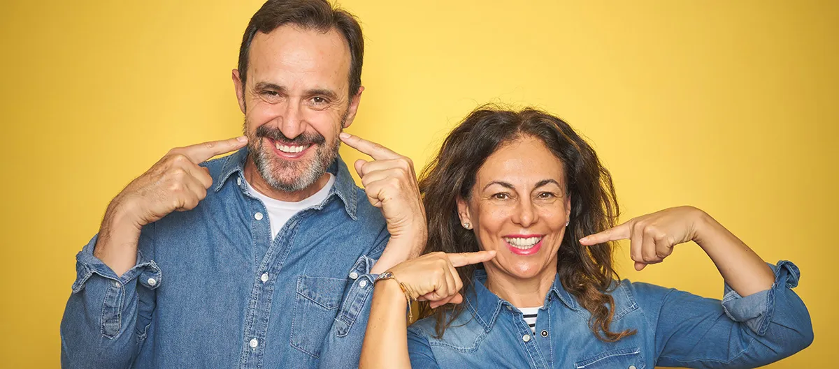 middle aged man and woman in denim shirts smiling and pointing at their new teeth.