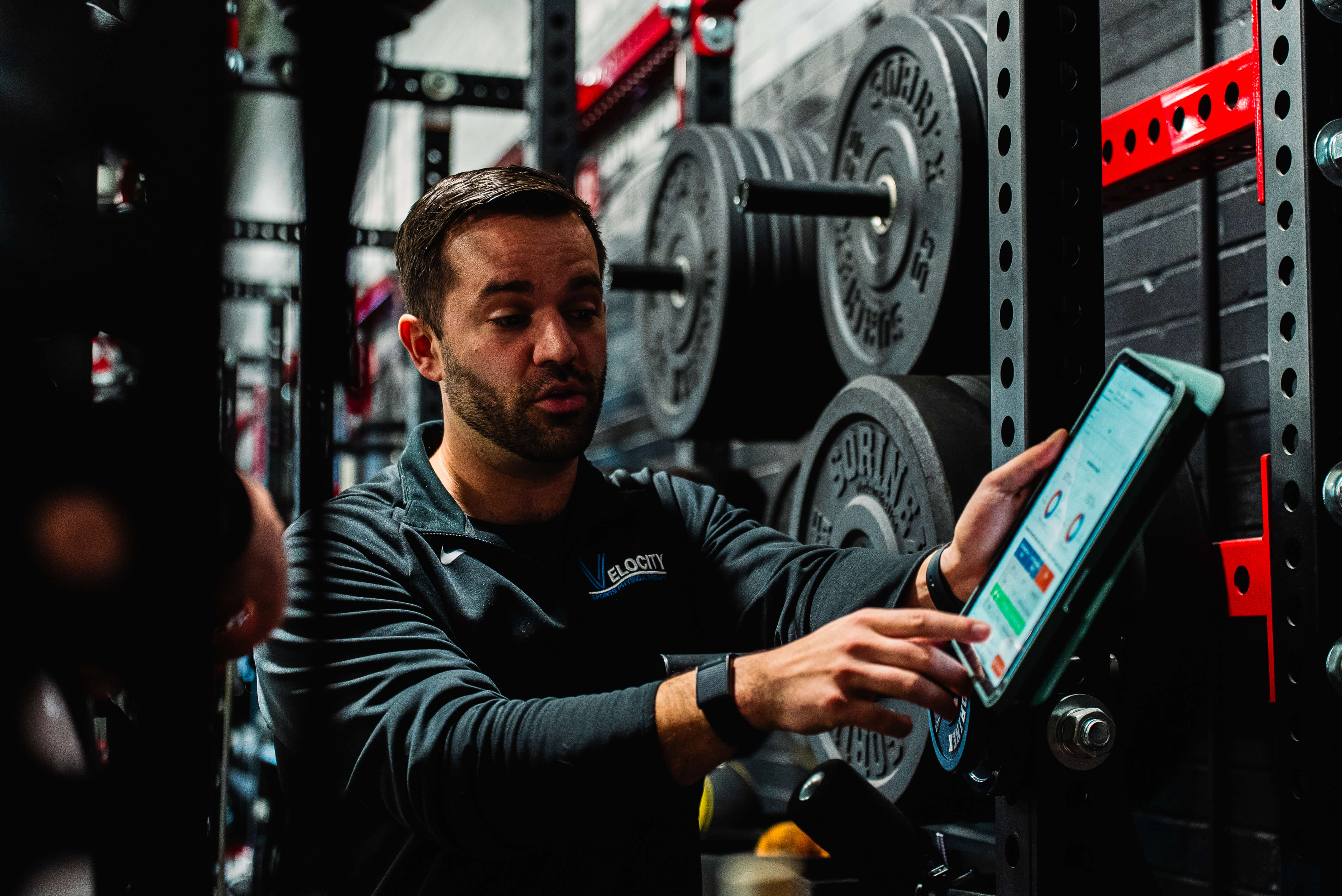 Man in a gym pointing at a tablet screen mounted on weightlifting equipment with stacked weight plates in the background.