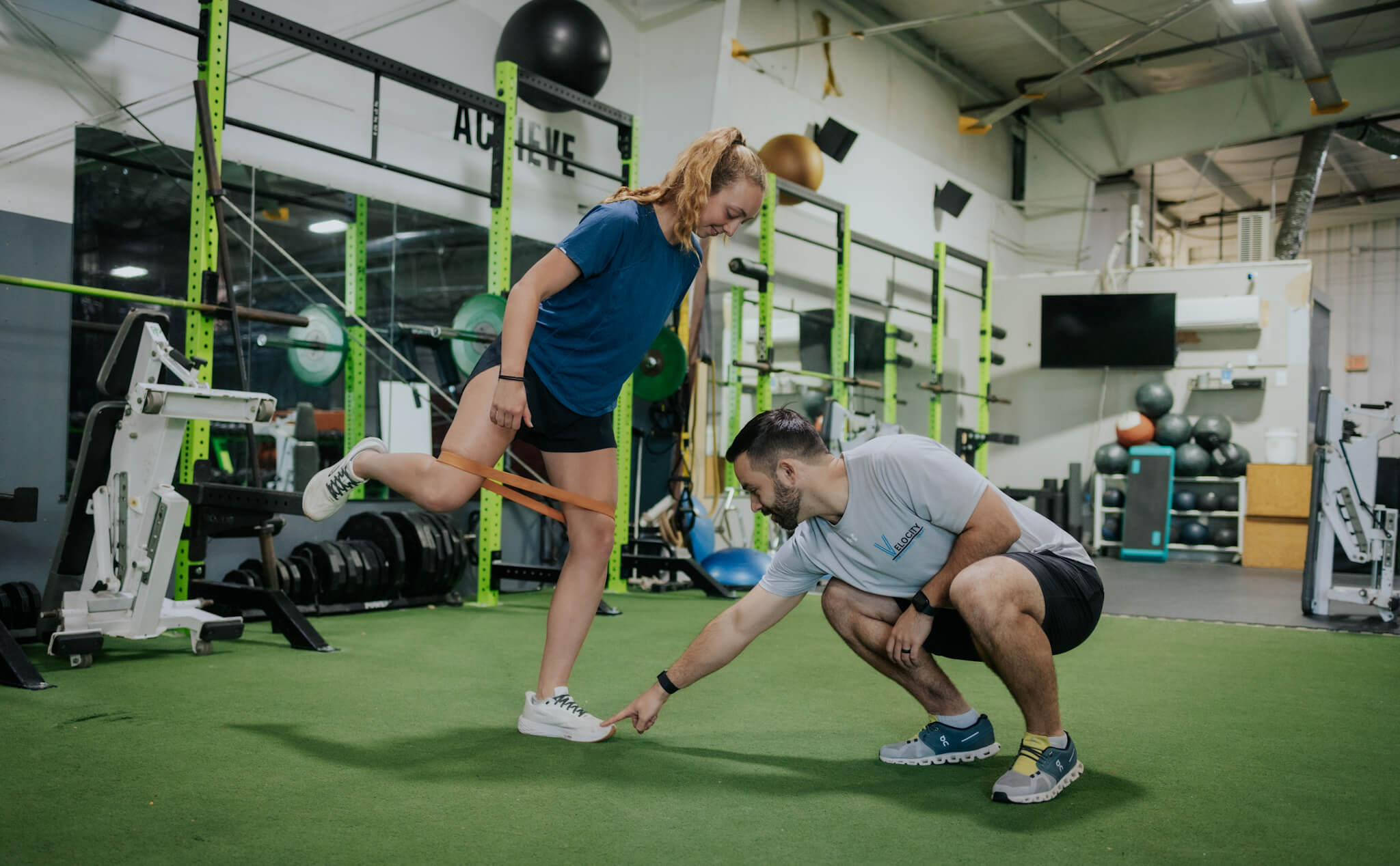 Woman performing resistance band leg exercise while trainer points to her foot in a gym with green turf flooring.