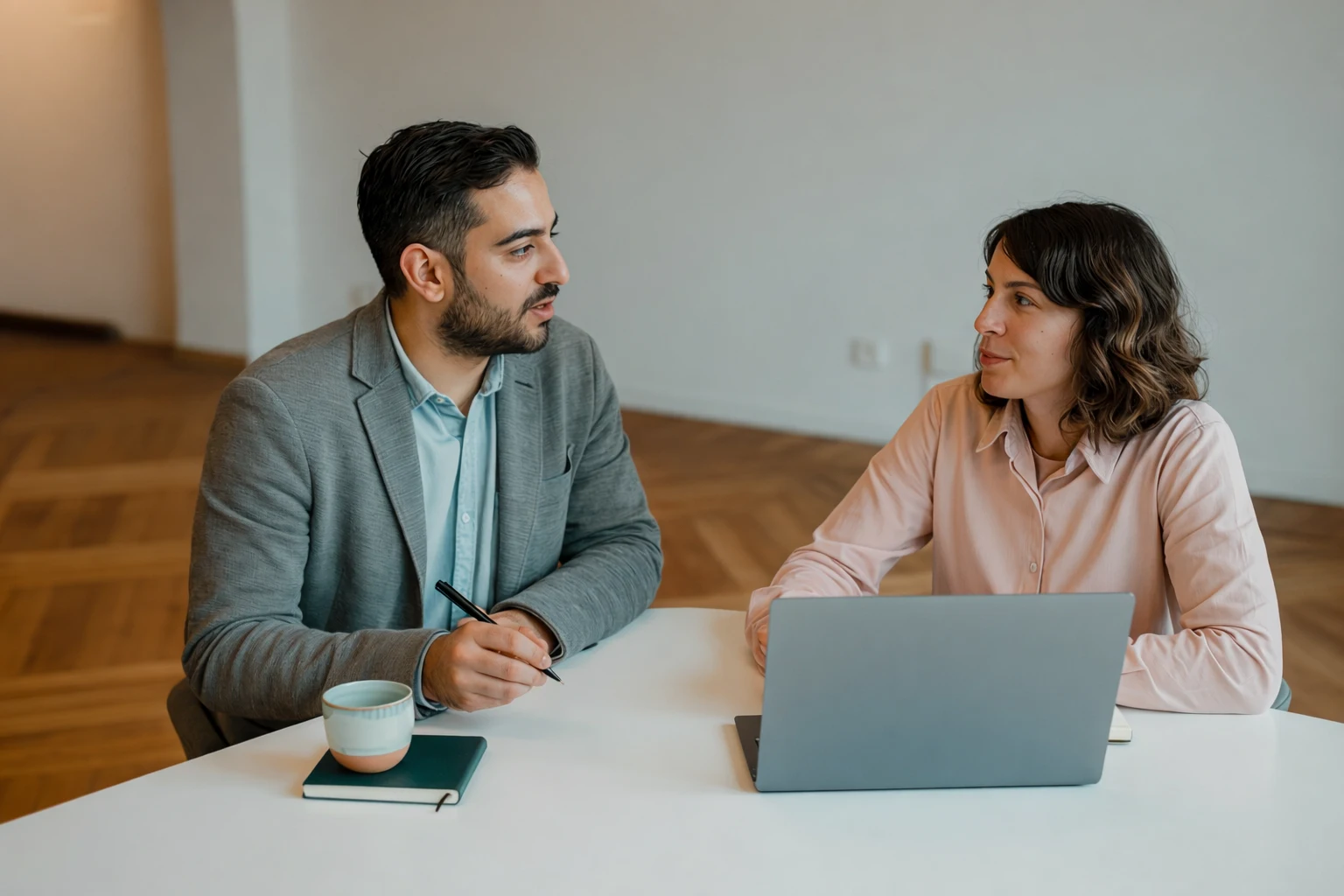 Man and woman having a conversation at a table with a laptop, notebook, and coffee cup.