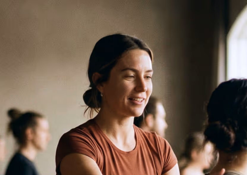Smiling woman in a brown shirt with tied-back hair, talking to another person indoors.