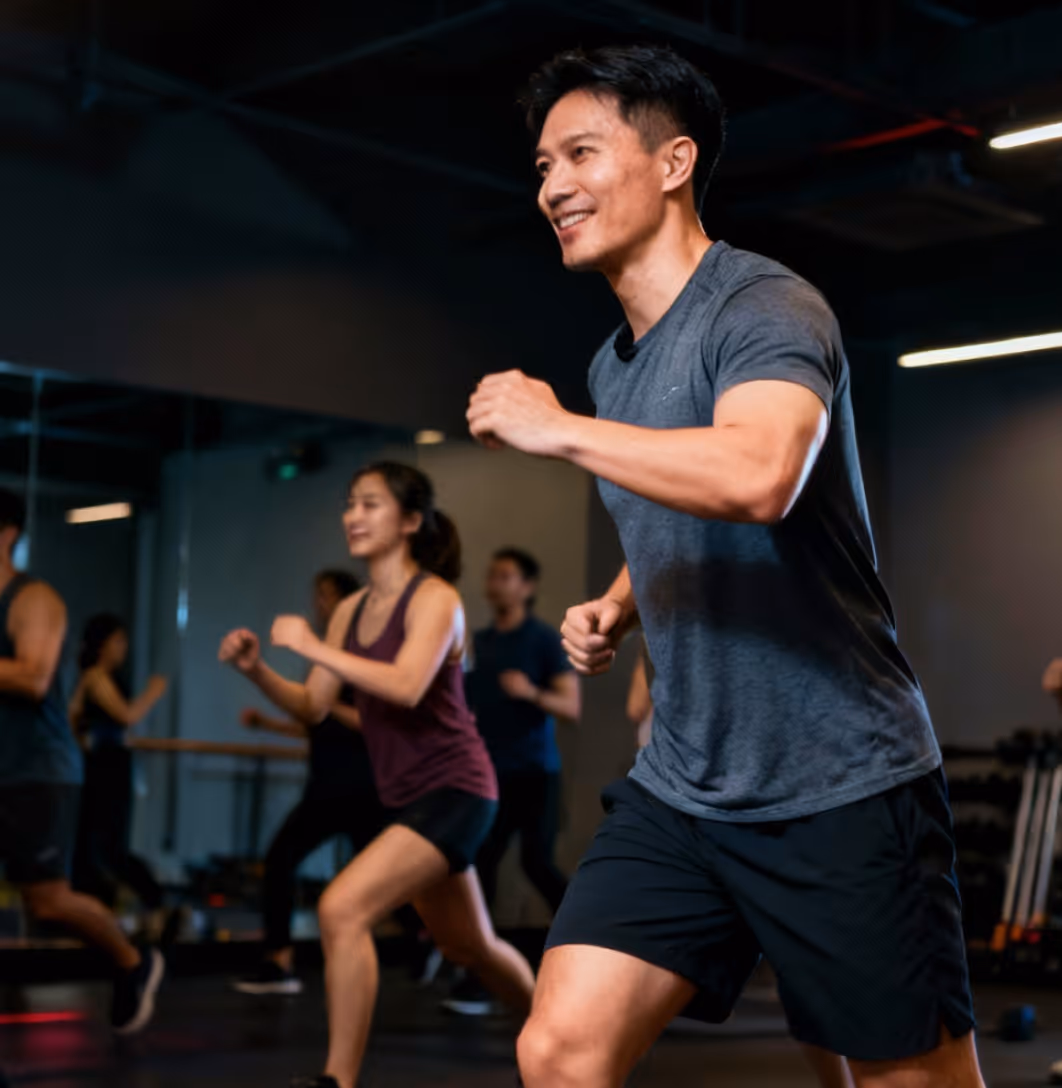 Group of people engaged in a lively fitness class indoors, with a smiling man in the foreground doing aerobic exercise.