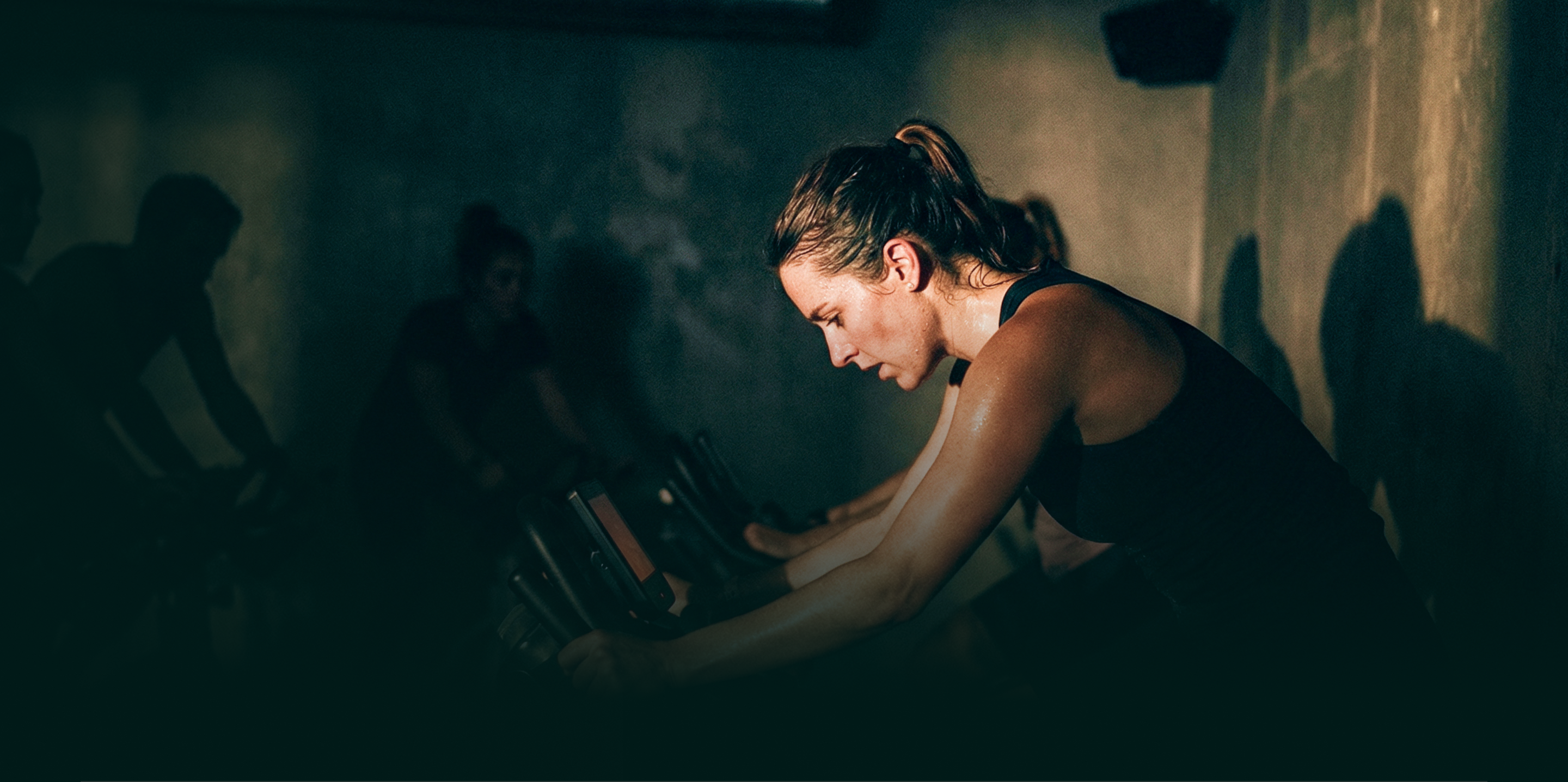 Woman intensely cycling on a stationary bike in a dimly lit indoor cycling class.