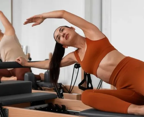 Woman in rust-colored activewear doing a side stretch on Pilates reformer equipment in a studio.