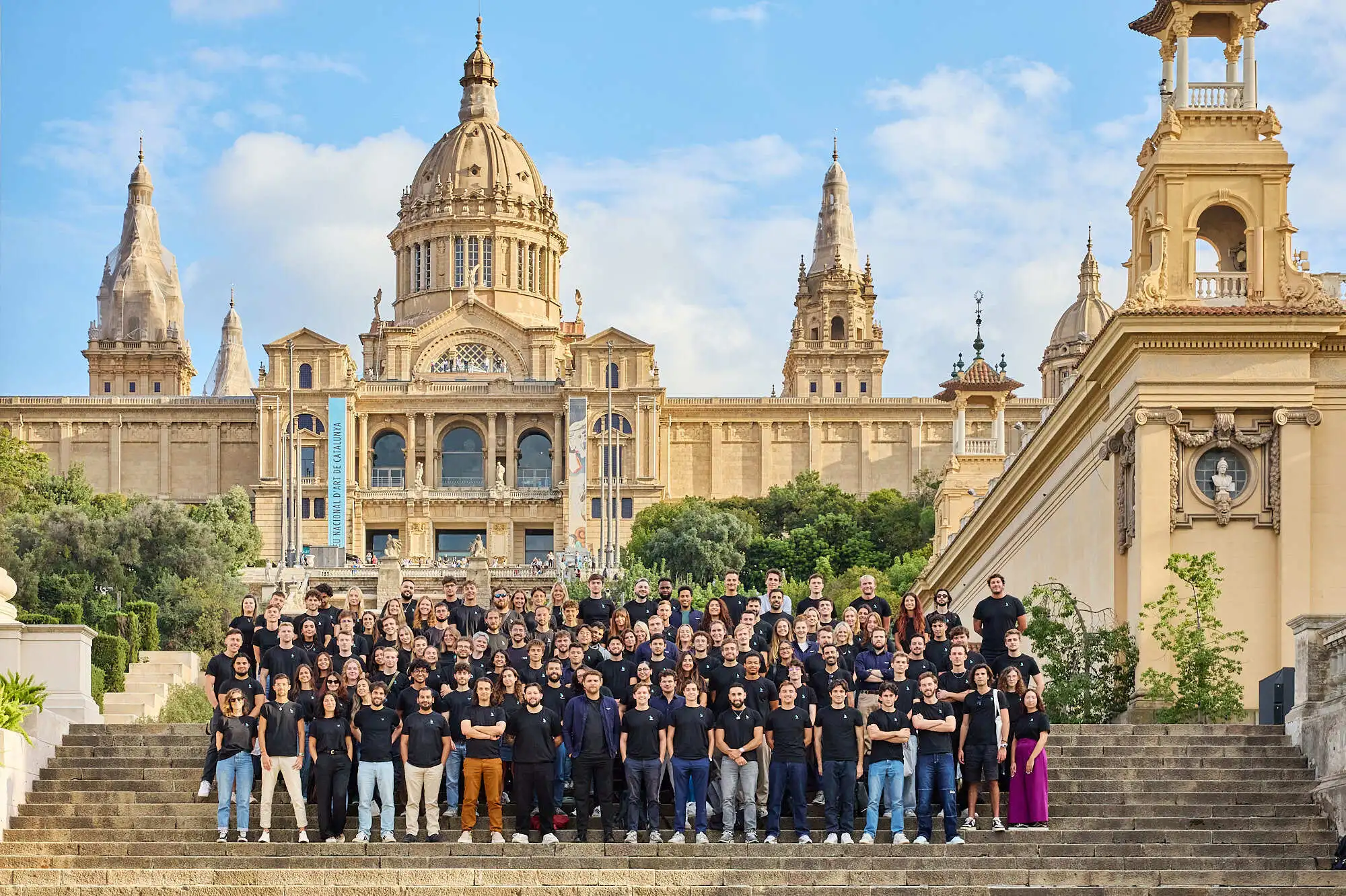 Large group of people wearing black shirts posing on stone steps with historic building in the background under a blue sky.
