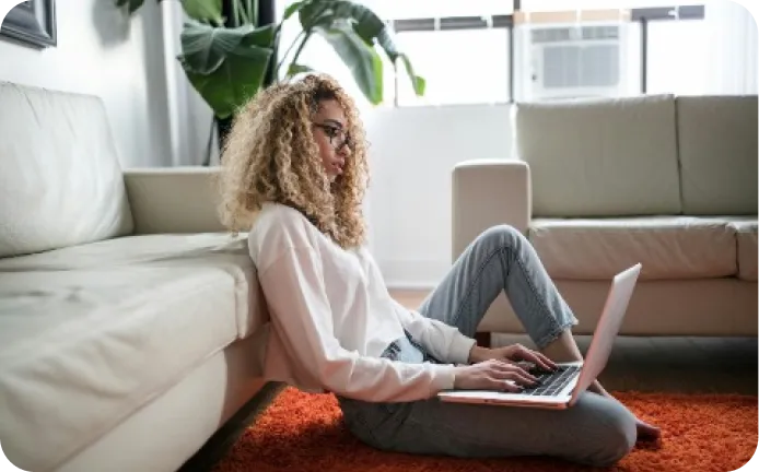 Woman with curly hair wearing glasses and a white sweater sitting on an orange carpet using a laptop in a bright living room.