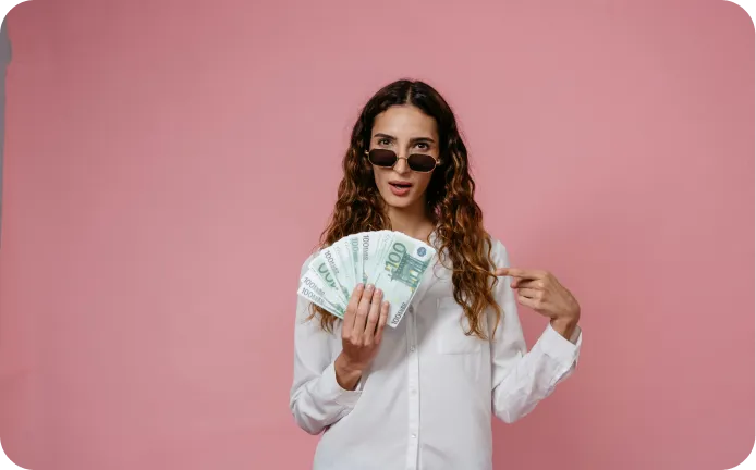 Woman wearing sunglasses and white shirt holding and pointing to several 100 euro banknotes against a pink background.