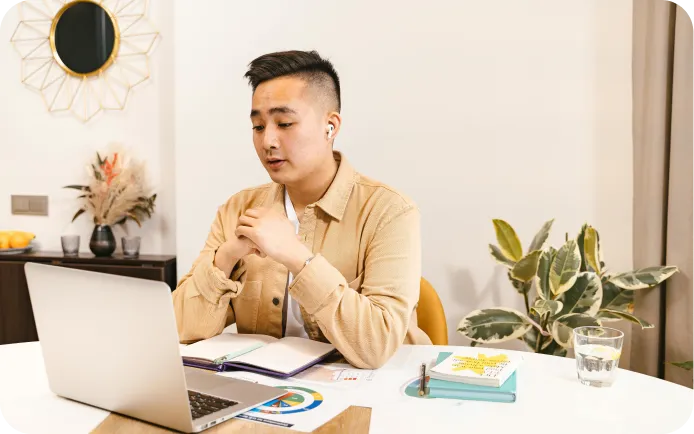 Young man with earbuds sitting at a desk, engaged in a video call on a laptop with notebooks and a glass of water nearby.