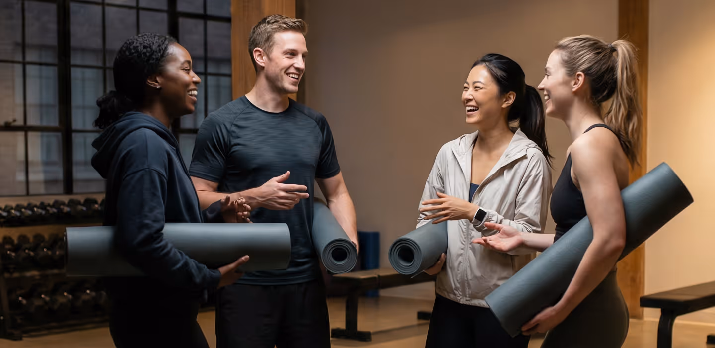 Four people holding yoga mats smiling and chatting in a gym setting.