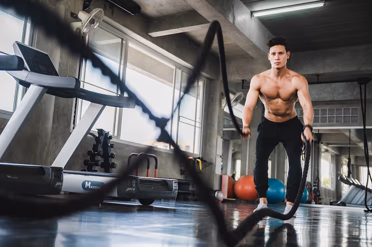 Un homme musclé, torse nu, s'entraînant avec des cordes d'entraînement dans une salle de sport moderne équipée de tapis de course et de ballons d'exercice.