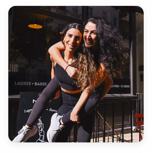 Two women in athletic wear smiling, one giving the other a piggyback ride outside a fitness studio.
