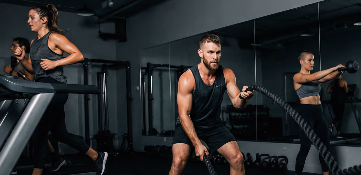 Three people exercising in a gym: a woman running on a treadmill, a man using battle ropes, and a woman lifting a kettlebell.