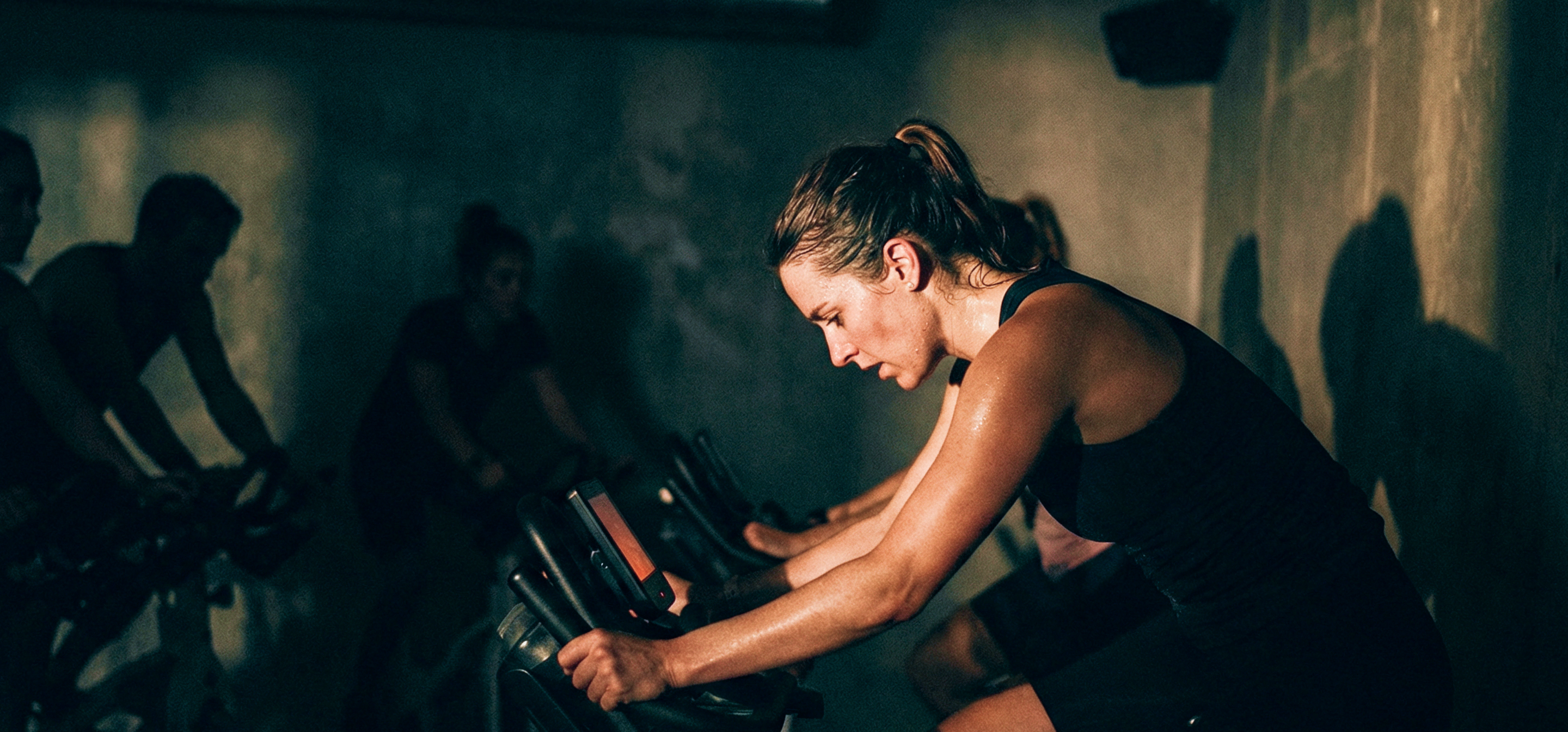 Sweating woman intensely cycling on a stationary bike in a dimly lit indoor cycling class.