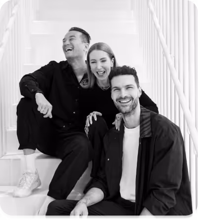 Tres adultos sonrientes sentados juntos en unas escaleras blancas: dos hombres y una mujer, vestidos de manera informal con ropa en blanco y negro.