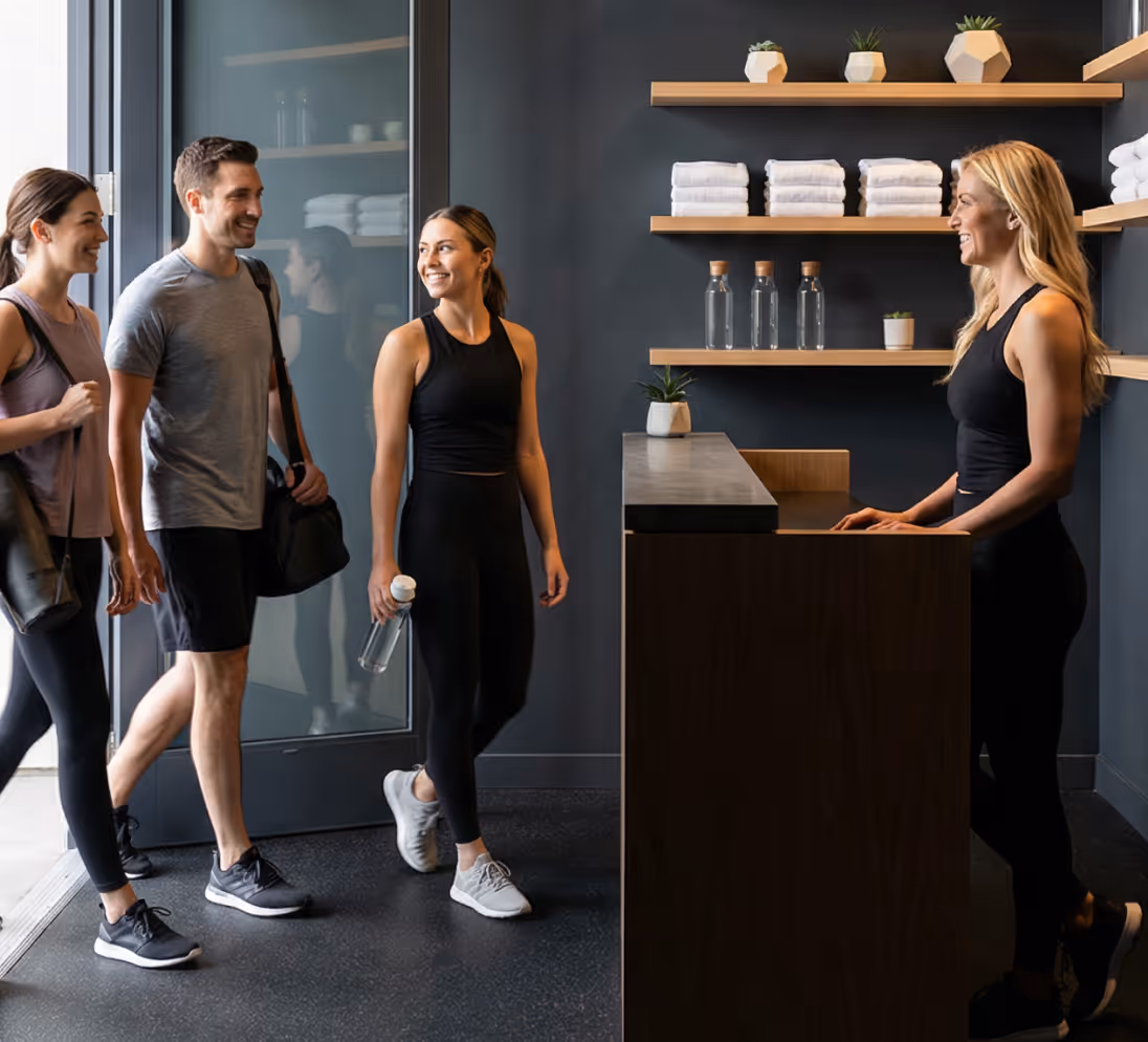 Three young people in athletic clothing entering a gym and greeting the smiling receptionist at the front desk.