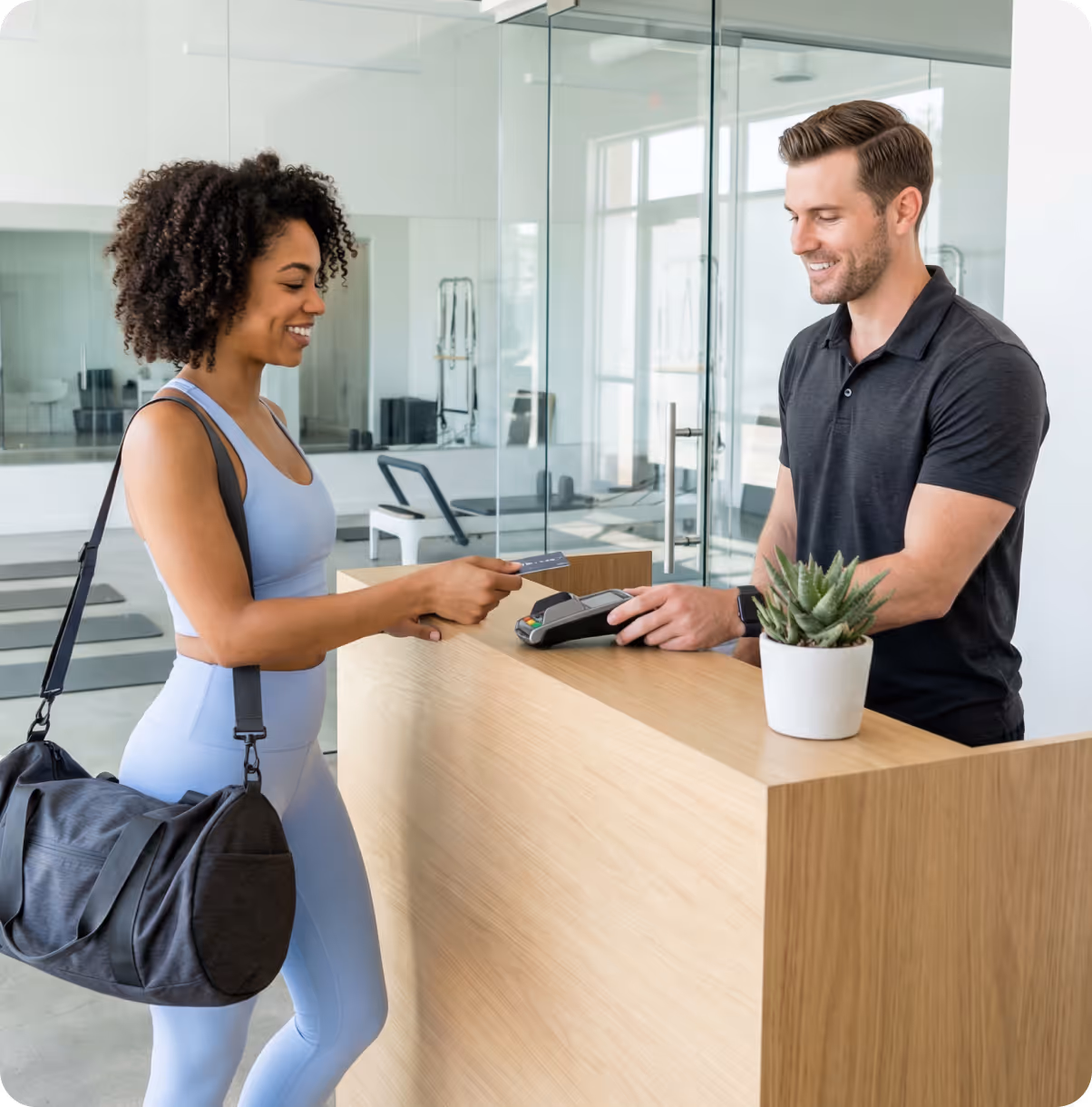 Smiling woman in activewear with a gym bag paying with a card at a gym reception desk attended by a smiling man.