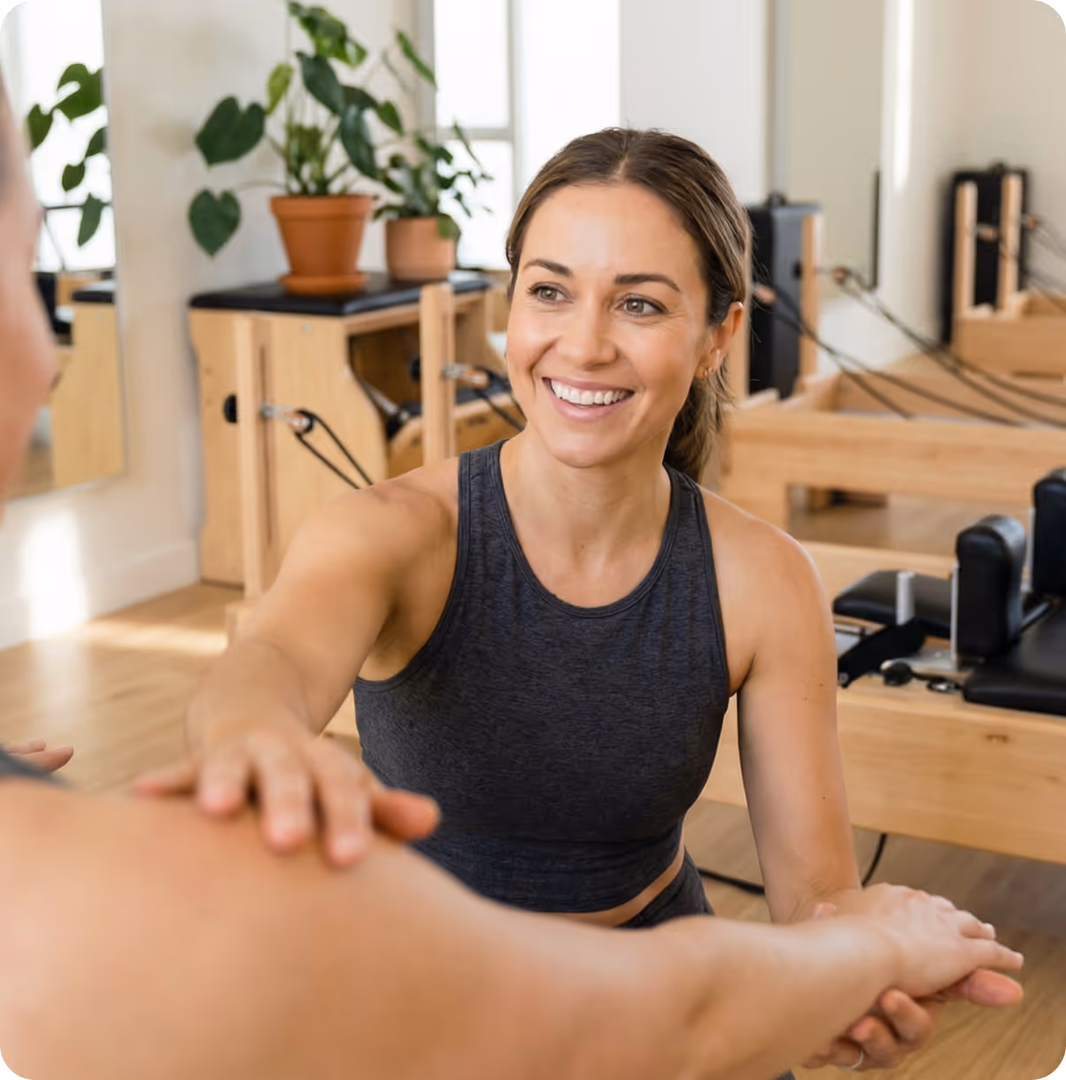 Smiling woman in workout attire guiding another person's arm stretch in a bright Pilates studio.