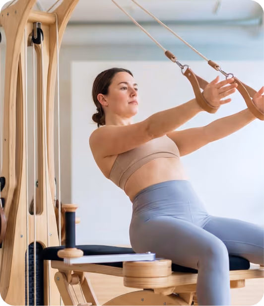 Woman exercising on a wooden Pilates reformer machine, pulling on straps with her arms extended.