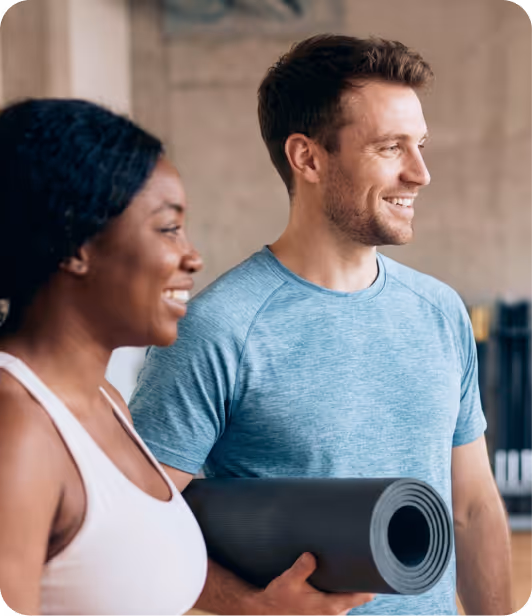 Smiling man in a blue shirt holding a rolled yoga mat standing next to a smiling woman in a white sports top.