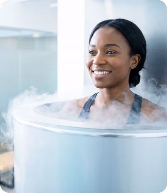 Smiling woman undergoing cryotherapy treatment inside a cylindrical chamber with mist.
