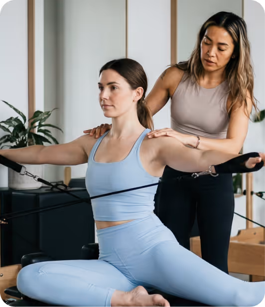 A Pilates instructor assists a woman in blue workout clothes performing a resistance exercise on Pilates equipment.