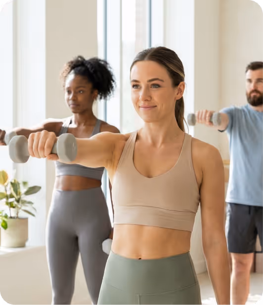 Three people in activewear performing barre exercises holding light dumbbells in a bright fitness studio.
