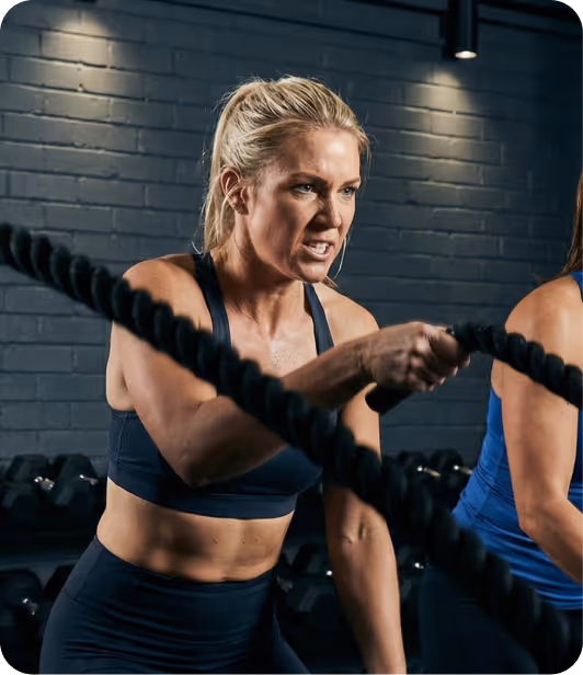 Determined woman exercising with battle ropes in a gym with brick wall background.