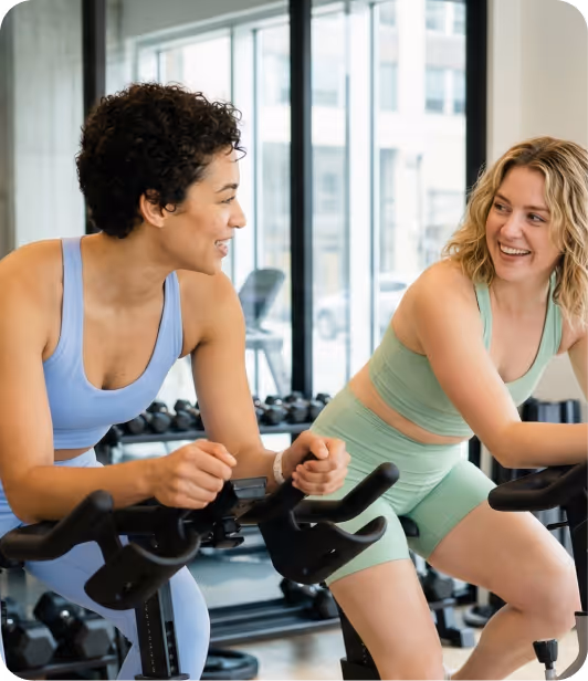 Two women smiling and talking while riding stationary bikes in a bright gym.