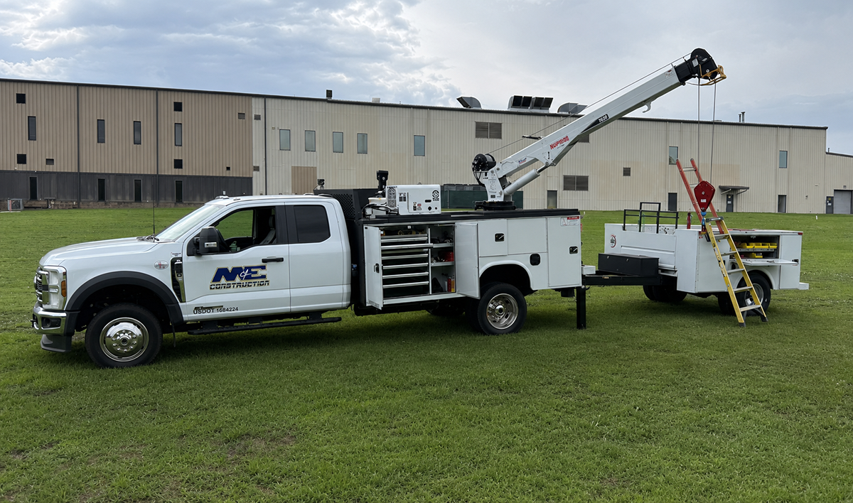 White construction service truck with an extended crane arm and open tool compartments, attached to a utility trailer with a yellow ladder and equipment, parked on green grass in front of a large industrial building under a cloudy sky.