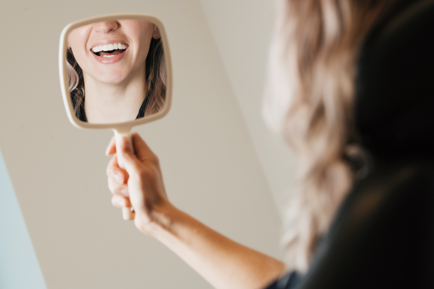 Woman smiling and looking at her reflection in a handheld mirror.