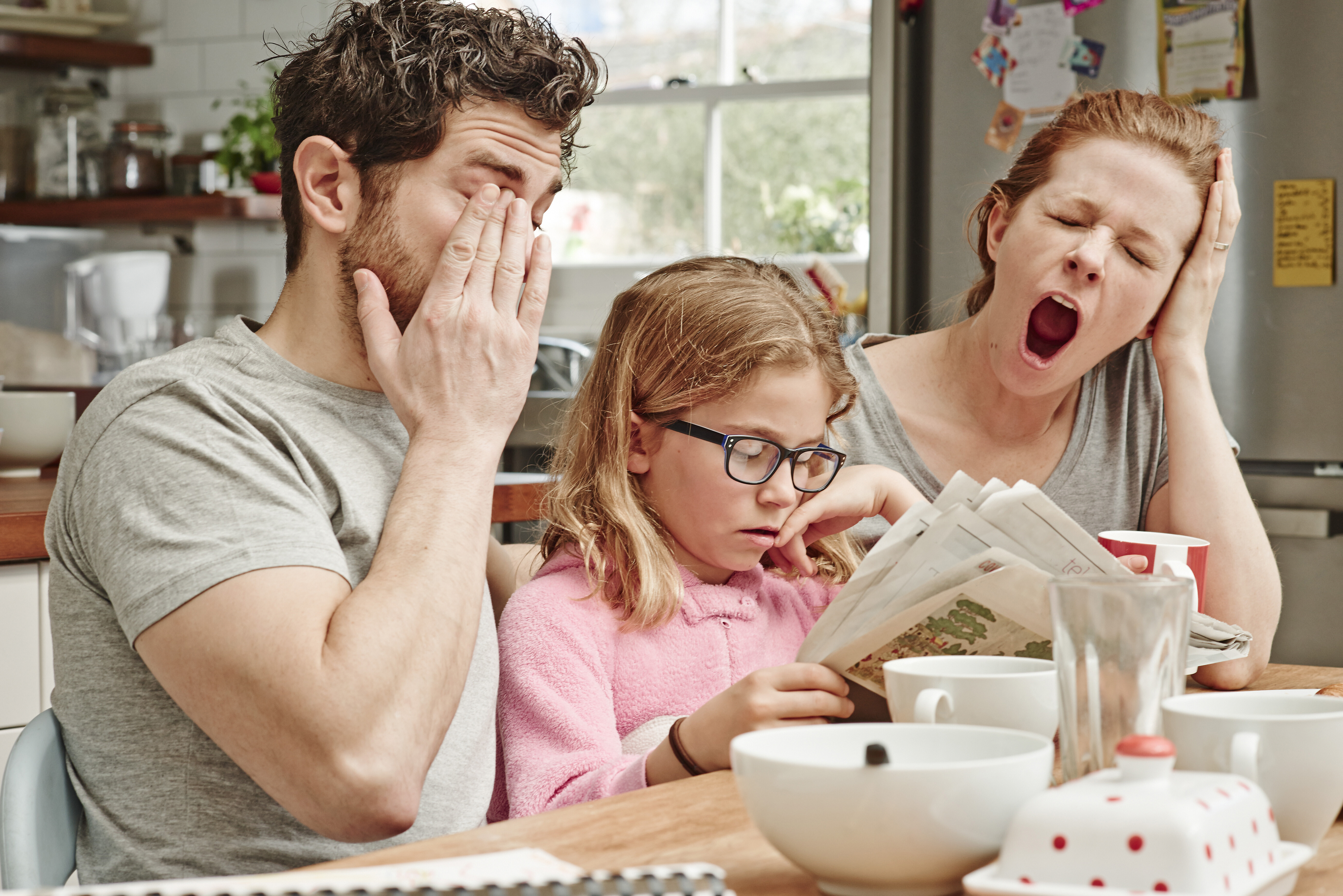 A tired family at the breakfast table, both parents yawning while their daughter reads