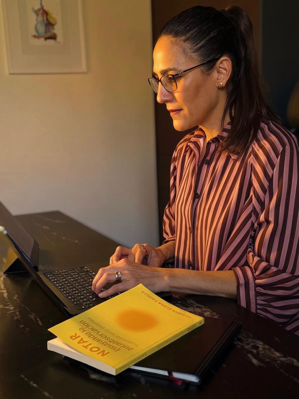 Woman in glasses and striped shirt typing on a laptop at a dark table with two books nearby.