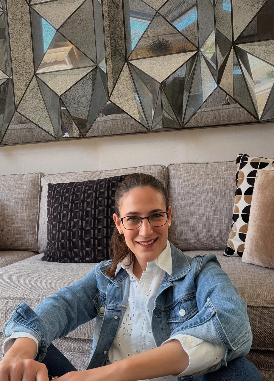 Smiling woman wearing glasses and a denim jacket sitting on a beige sofa with decorative cushions and a geometric mirror behind her.