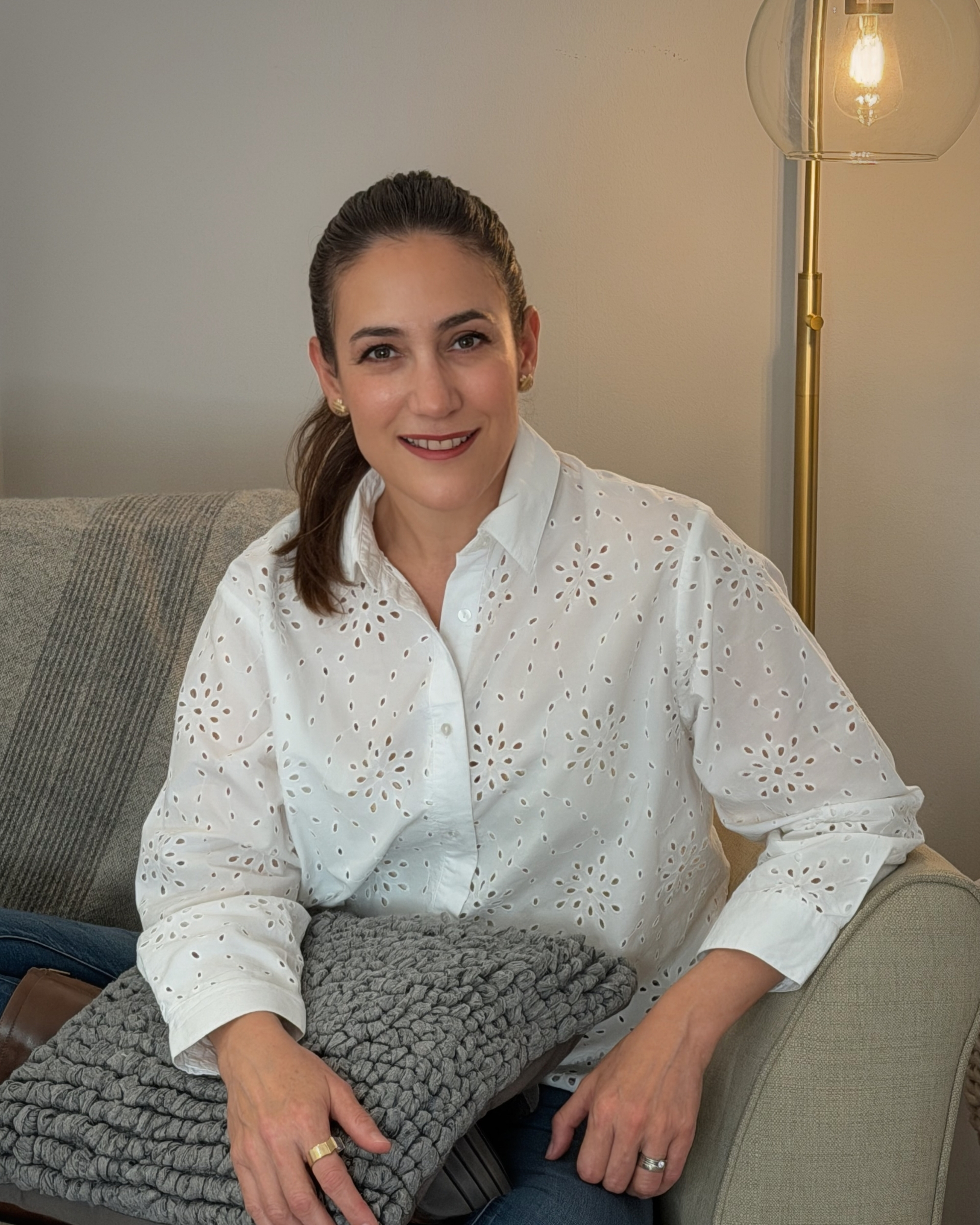 Claudia Padilla Smiling woman sitting on a sofa holding a textured gray pillow, wearing a white eyelet blouse and blue jeans.