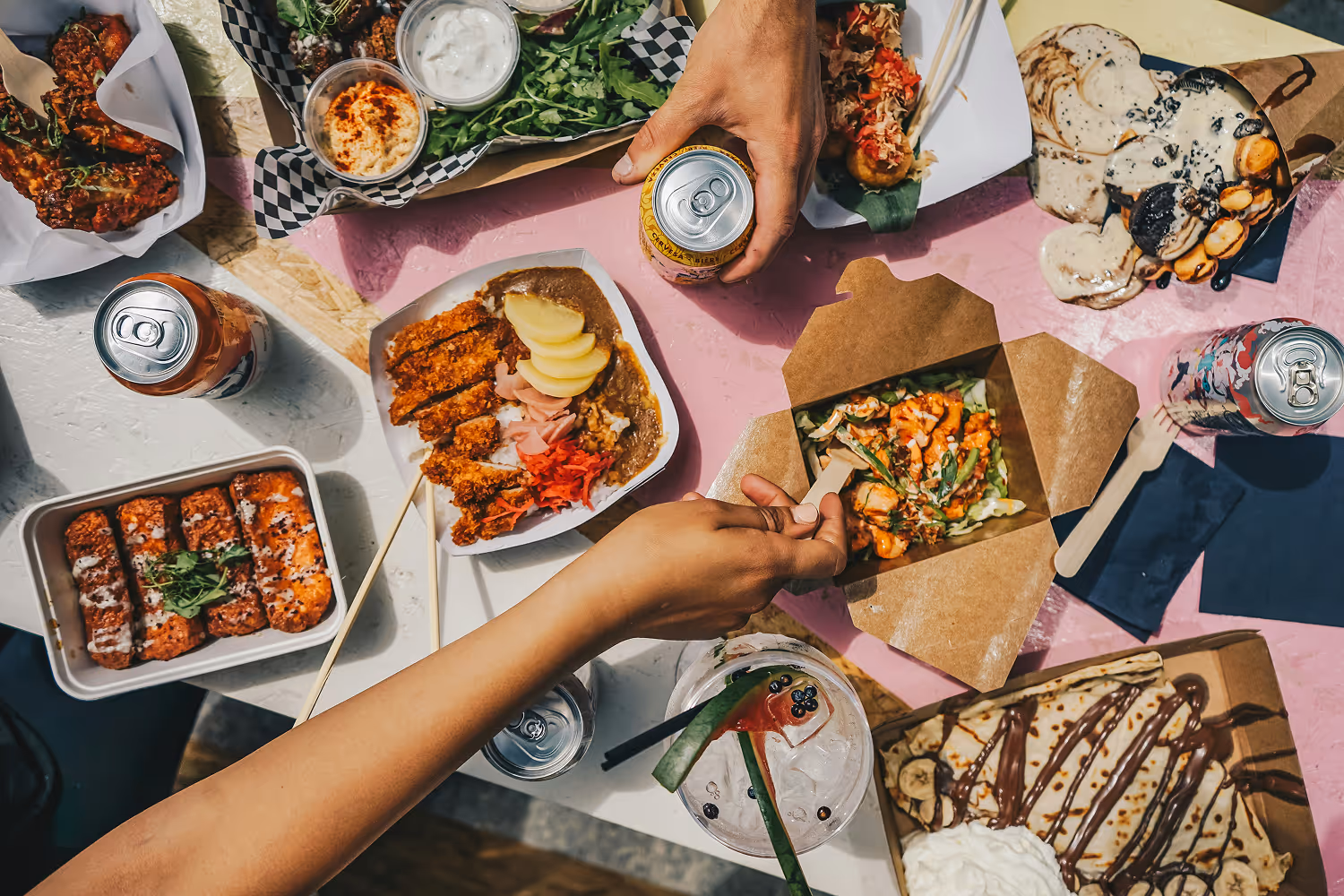 Overhead view of a table set with various Asian-inspired takeout dishes, including sushi, curry with rice, salad, and desserts, with two hands holding a can and a wooden fork.