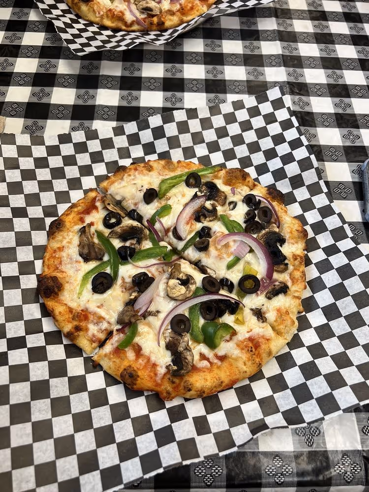 Small vegetable pizza with black olives, green peppers, mushrooms, and red onions on checkered paper.