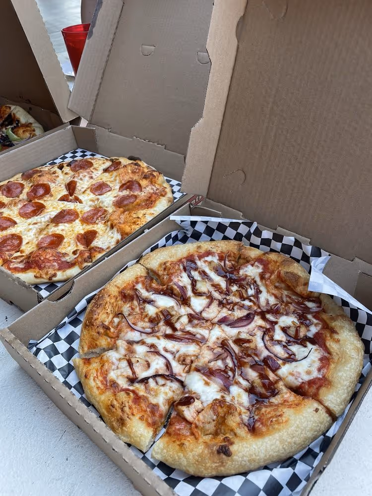 Two open pizza boxes on a white surface, one with a pepperoni pizza and the other with a chicken and red onion pizza drizzled with barbecue sauce.
