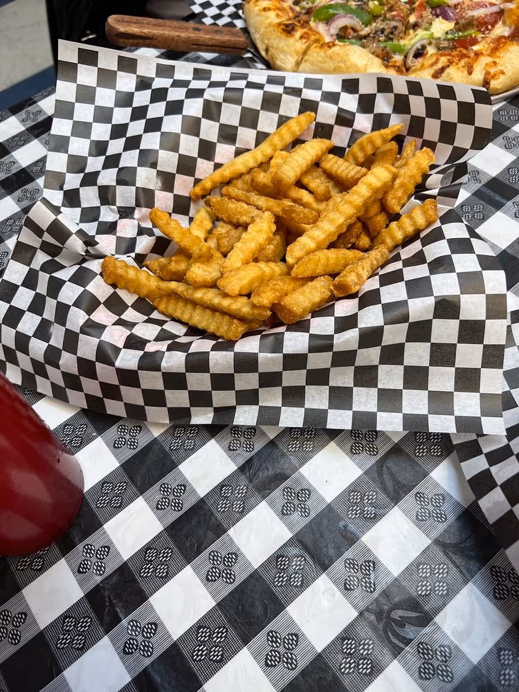 Crinkle-cut French fries served in a black and white checkered paper basket with a pizza slice partially visible in the background.