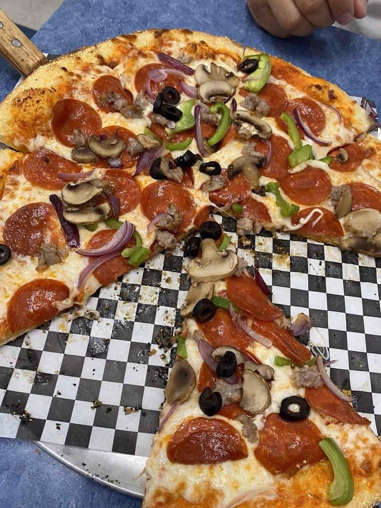 Pepperoni pizza topped with mushrooms, green bell peppers, red onions, black olives, and sausage, with one slice separated on a black-and-white checkered paper-lined tray.