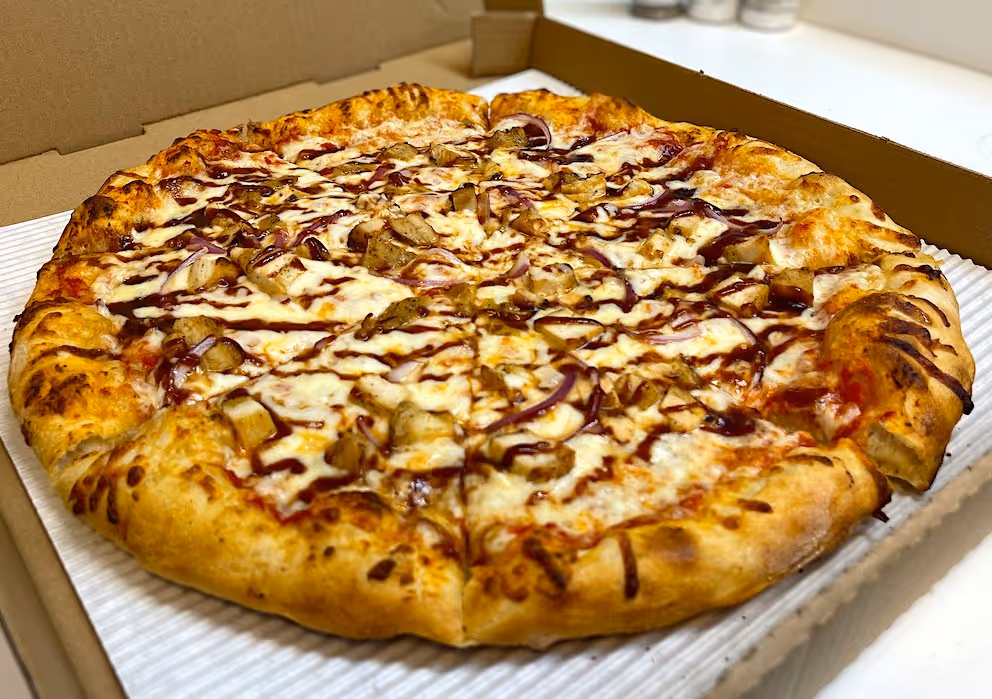 Freshly baked pizza topped with melted cheese, grilled chicken pieces, red onion slices, and barbecue sauce in a cardboard delivery box.