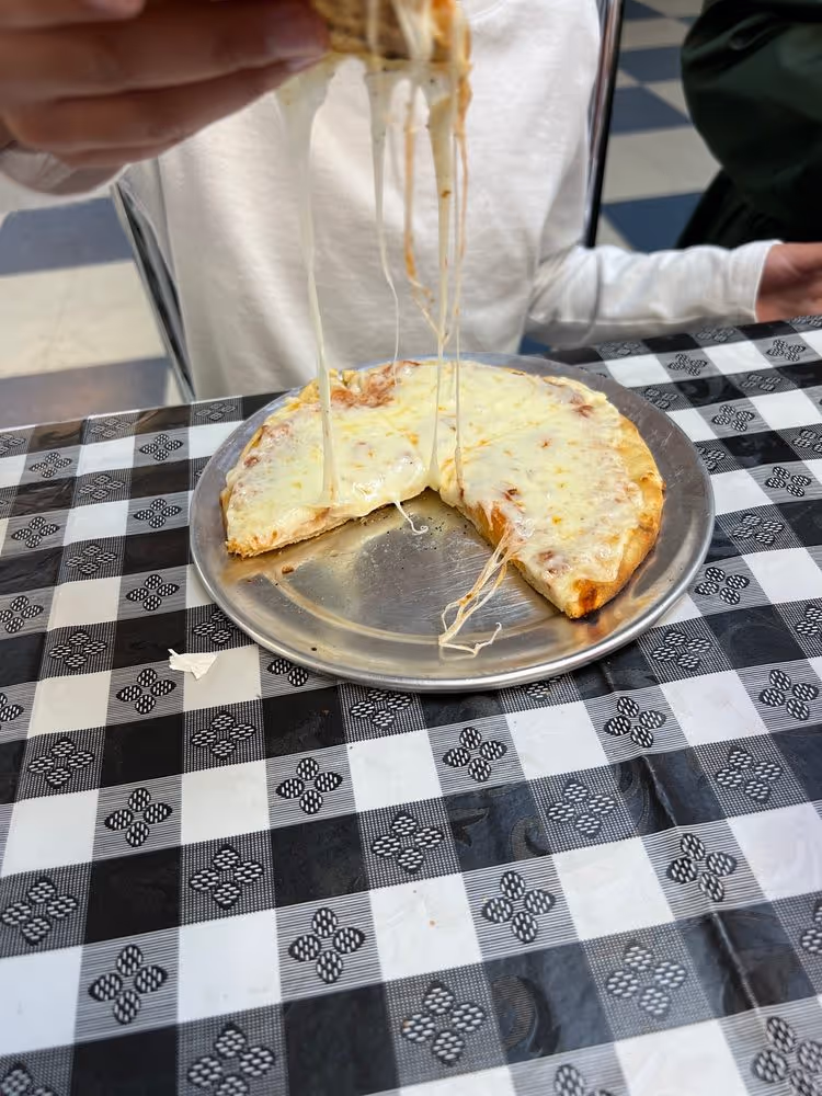 Person lifting a slice of cheese pizza with melted cheese stretch on a metal tray over black and white checkered tablecloth.
