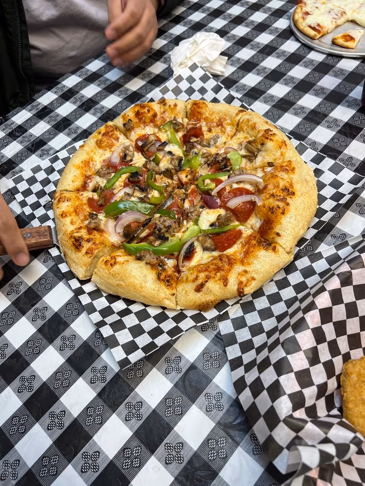 Small pizza with green bell peppers, onions, mushrooms, and pepperoni on a checkered paper-lined table.