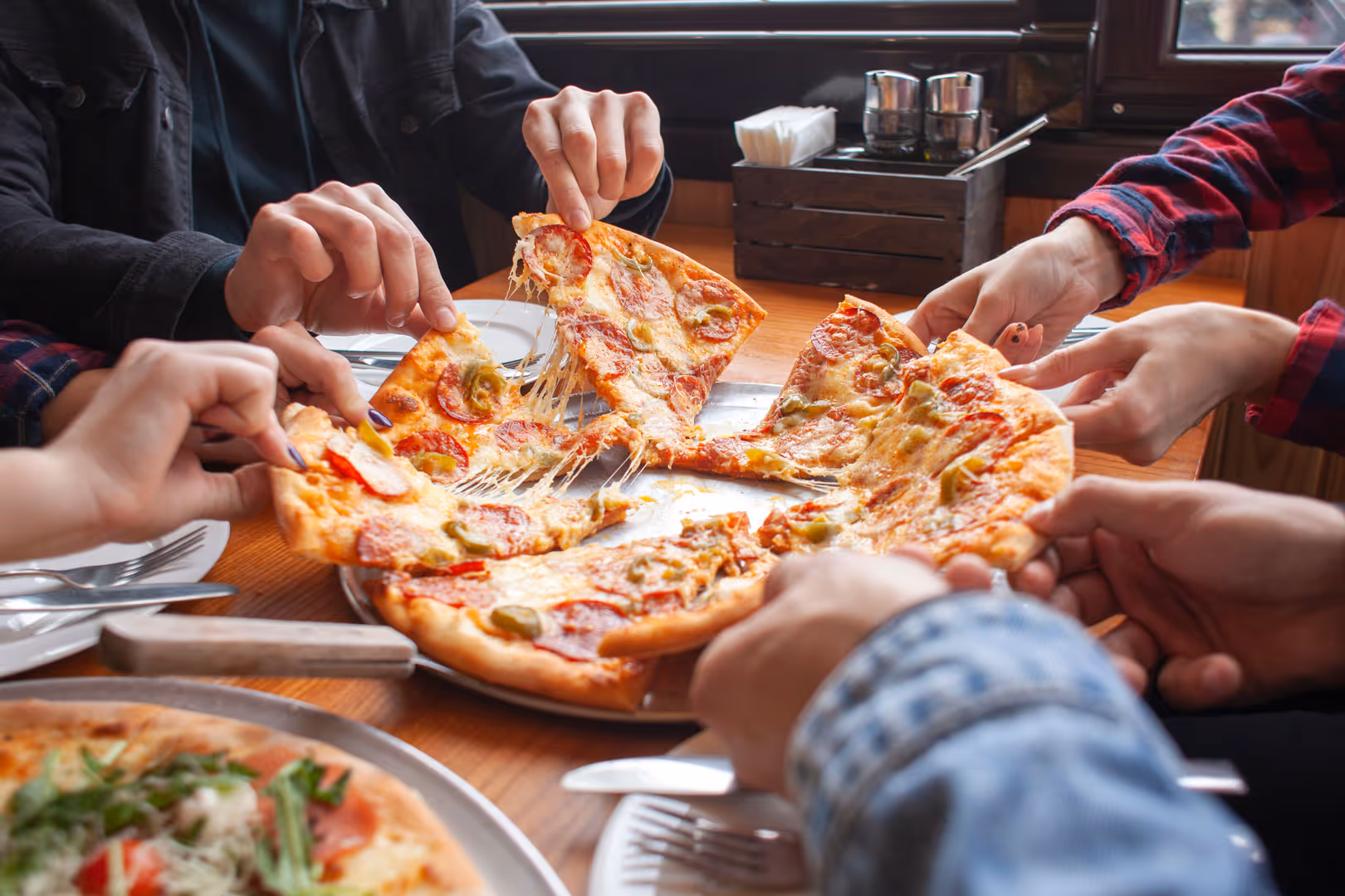 Multiple hands reaching to grab slices of pepperoni and cheese pizza from a metal tray at a wooden table.