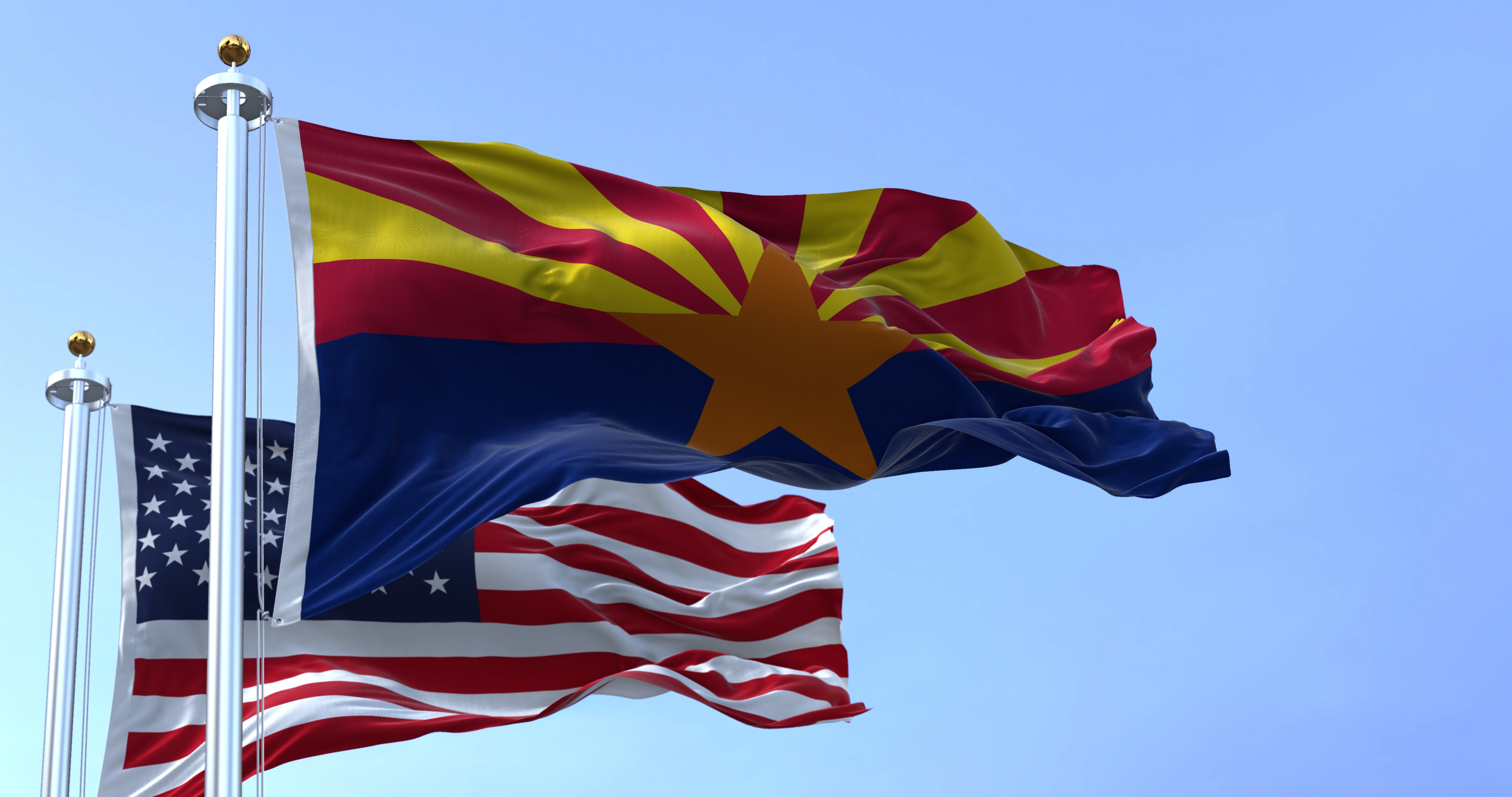 Arizona state flag with a large copper star and alternating red and yellow rays flying next to the United States flag against a clear blue sky.